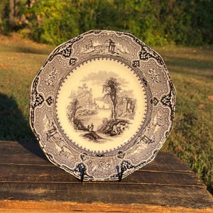 May include: A ceramic plate with a scalloped edge featuring a black and white transferware print. The center depicts a landscape with ruins and trees, while the border displays intricate floral patterns and architectural motifs.
