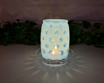 May include: A cylindrical white wax warmer with a repeating blue polka dot pattern, featuring a lit tea light candle inside. The warmer sits on a clear, scalloped glass dish that reflects the warm light outward in a radial pattern.