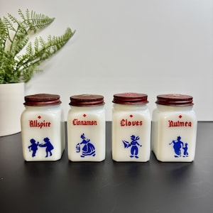 May include: Set of four vintage spice jars with white glass bodies and red metal lids. Each jar is labeled with a spice name in red script and features a blue illustration. Spices include Allspice, Cinnamon, Cloves, and Nutmeg.