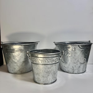 May include: Three galvanized metal buckets of varying sizes. The buckets have a silver finish and feature handles. The middle bucket has a decorative floral design around the top. The buckets are suitable for indoor or outdoor use.