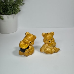 May include: A pair of ceramic bear figurines, one holding a black and yellow pot, the other with its paw near its mouth. The bears are light brown with textured fur details. These decorative figurines are likely salt and pepper shakers.