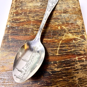 May include: A silver spoon with an engraved design. The spoon's bowl features a detailed depiction of a ship, with the words "GENERAL GUTHRIE" and "SOUTH GUTHRIE" inscribed. The handle has decorative elements and text. The spoon is set against a weathered wooden surface.