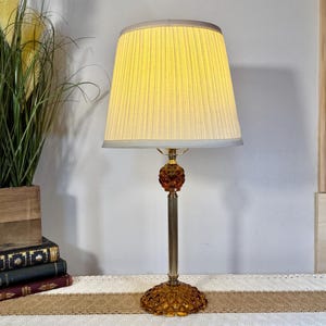 May include: A vintage amber glass table lamp with a pleated lampshade. The lamp features a round base and a decorative orb in the center. The lampshade is a light yellow color. The lamp is approximately 24 inches tall.