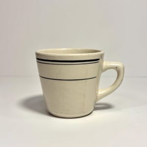 May include: A cream-colored ceramic mug featuring a tapered body and a side handle. The exterior is decorated with two thin, horizontal black lines encircling the upper portion of the mug, providing a simple contrast against the light base color.