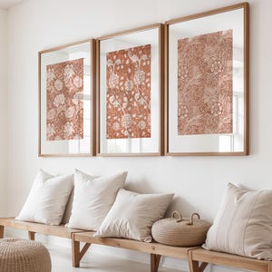 May include: Three framed floral art prints in terracotta and cream tones are displayed above a wooden bench with white cushions. The prints showcase intricate botanical designs, enhancing the room's decor. A woven basket and a striped cushion complete the scene, adding a touch of elegance.