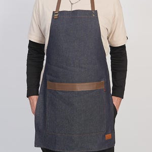 Puede incluir: Un delantal de mezclilla azul oscuro con correas de cuero marrón para el cuello y la cintura, herrajes metálicos y un bolsillo frontal grande con borde de cuero marrón. El delantal incluye un pequeño parche rectangular marrón en la esquina inferior.