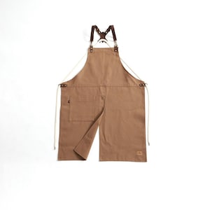 以下が含まれることがあります： 胸ポケットとスプリットフロントデザインのタンカラーのエプロン。背中で交差する茶色の革ストラップと、金属製の金具でエプロンに取り付けられています。ウエストタイには白いコードが使用されています。