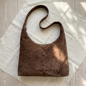Puede incluir: Un bolso hobo de ante marrón oscuro con una sola correa para el hombro y una costura central vertical decorada con tres detalles de cordones de cuero entrecruzados. El bolso tiene una silueta suave y holgada con una textura mate uniforme.