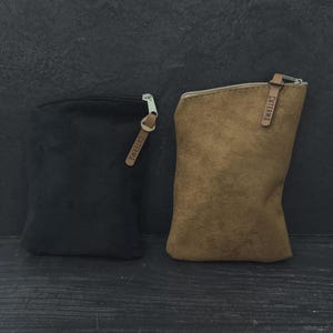 May include: Two small, rectangular pouches. One is black, the other is brown. Both have a zipper closure with a silver pull and a leather tag that reads "CUTERMA". The pouches are made of a soft material.