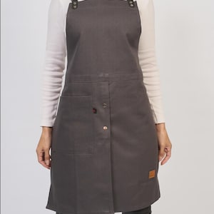 Puede incluir: Un delantal gris carbón que presenta un bolsillo frontal rectangular, una fila vertical de remaches metálicos y correas ajustables para los hombros sujetas con herrajes de metal plateado. Un pequeño parche rectangular marrón es visible.