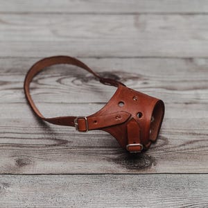 Puede incluir: Un bozal de perro de cuero marrón con correa y cierre de hebilla. El bozal tiene varios agujeros pequeños y está diseñado para adaptarse al hocico de un perro. El cuero es de un color marrón cálido y rico.