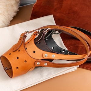 May include: A collection of leather dog muzzles in various colors, including tan, black, and brown. The muzzles have adjustable straps and are designed with multiple ventilation holes. They are displayed on a white cloth and a brown surface.