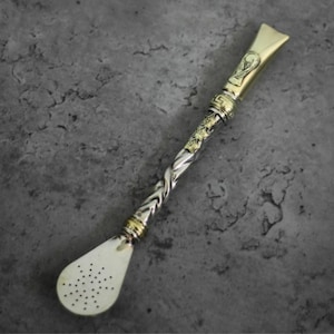 Puede incluir: Una bombilla de yerba mate plateada y dorada con un tallo retorcido y un filtro en forma de cuchara. La parte superior presenta un diseño dorado y el filtro tiene pequeños agujeros. La bombilla está diseñada para beber yerba mate.