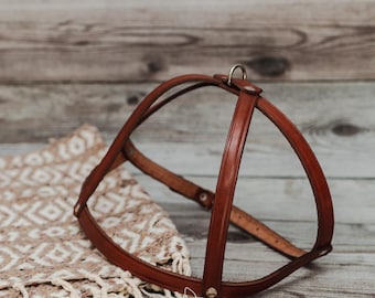 May include: A brown leather dog harness with a metal ring and buckle. The harness is displayed on a wooden surface with a patterned textile. The harness is designed to fit around the dog's chest and back.