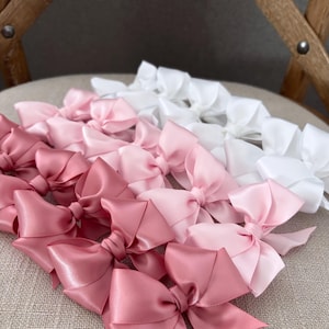 May include: A collection of satin ribbon bows in shades of pink and white. The bows are arranged in a gradient from white to pink, with the darkest pink bows at the bottom of the image.
