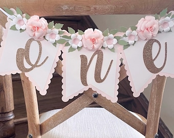 May include: A decorative banner featuring three white scalloped pennants with light pink backing. Each pennant displays a large, glittery copper-colored letter spelling out the word one. The top is adorned with paper flowers in shades of pale pink and white, accented with sage green paper leaves.