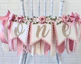 May include: A decorative banner featuring three white pennants with scalloped edges, each displaying a gold glitter letter spelling out the word one. The banner is adorned with pink paper flowers, green leaves, and long pink and white ribbons.