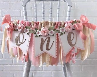 Puede incluir: Una guirnalda para trona con tres banderines blancos de bordes festoneados y letras cursivas de purpurina color cobre que forman la palabra one. La guirnalda está decorada con rosas de tela rosa, hojas verdes y cintas en tonos rosa y crema.