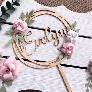 Peut inclure: Une décoration de gâteau en bois composée d'un cadre circulaire avec le prénom Evelyn écrit en lettres cursives. L'ornement est agrémenté de fleurs en papier en relief dans des tons rose pâle et blanc, avec des feuilles vertes et une tige.