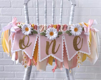 May include: A pink and gold "ONE" banner with daisy accents is draped over a white wooden chair. The banner has scalloped edges and gold glitter lettering. Ribbons in shades of pink, yellow, and cream hang below the banner.