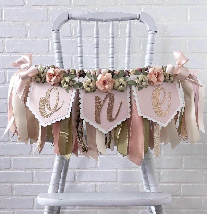 K&ouml;nnte beinhalten: Ein dekoratives Hochstuhl-Banner mit drei hellrosa Wimpeln mit gewelltem Rand, die jeweils einen glitzernden goldenen Buchstaben tragen, um das Wort one zu bilden. Das Banner ist mit k&uuml;nstlichen blassrosa Rosen und Stoffb&auml;ndern verziert.