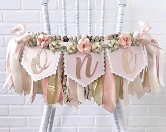 Peut inclure: Une bannière décorative pour chaise haute composée de trois fanions rose pâle avec le mot ONE en écriture dorée scintillante. La bannière est ornée de fleurs artificielles rose poudré et crème ainsi que de longs rubans roses, crème et dorés.
