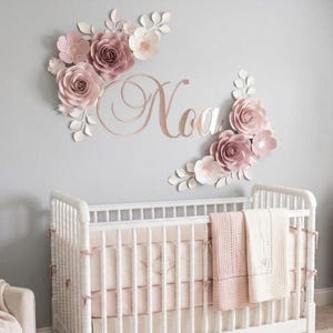 May include: A white wooden cot with a light pink blanket and a wall display featuring the name Noa in rose gold script lettering, surrounded by large paper flowers in shades of dusty rose and cream with white paper leaves and stems.