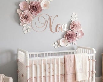 May include: A white wooden crib with a light pink blanket, featuring a wall display of large dusty rose and white paper flowers arranged around a metallic rose gold script sign that reads Noa in elegant cursive lettering.