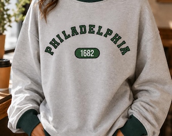 May include: A light gray long-sleeve sweatshirt featuring a dark green mock neck collar, dark green sleeve cuffs, and a dark green hem. The chest displays the text PHILADELPHIA in a dark green arched font above a dark green oval containing 1682.