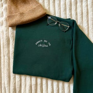 May include: A dark green sweatshirt with the text "Sundays are for The Birds" in white script. A tan beanie and silver-framed glasses are placed on the folded sweatshirt. The items are displayed on a textured, off-white surface.