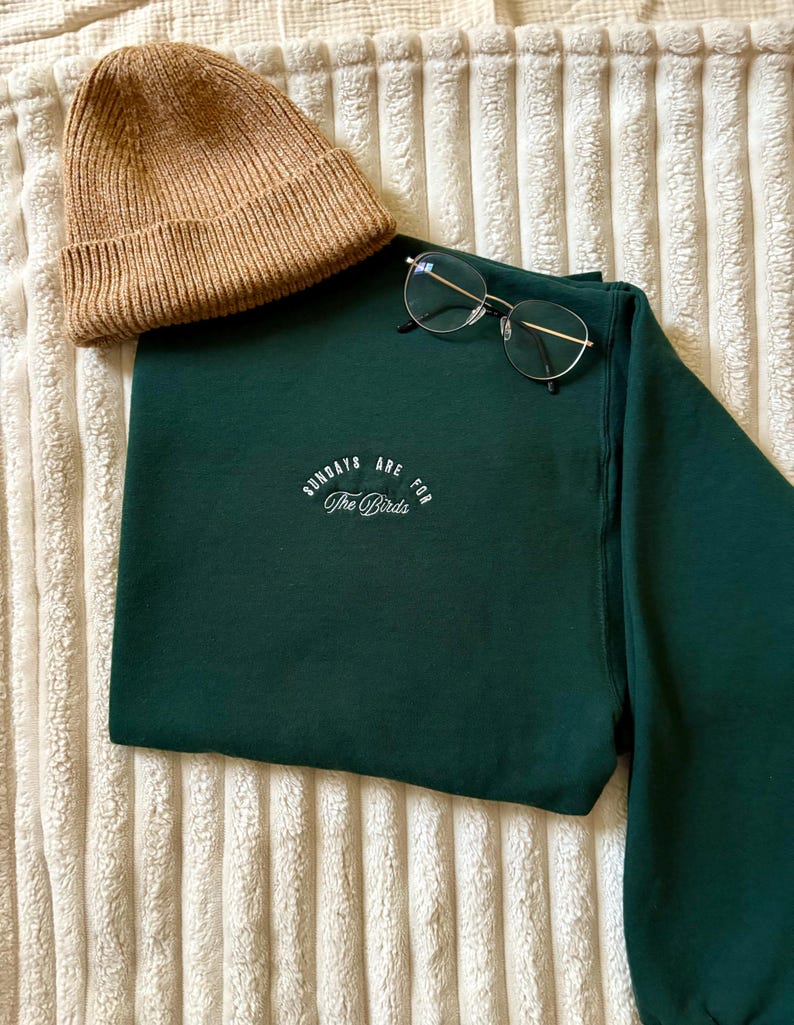 May include: A dark green sweatshirt with the text "Sundays are for The Birds" in white script. A tan beanie and silver-framed glasses are placed on the folded sweatshirt. The items are displayed on a textured, off-white surface.