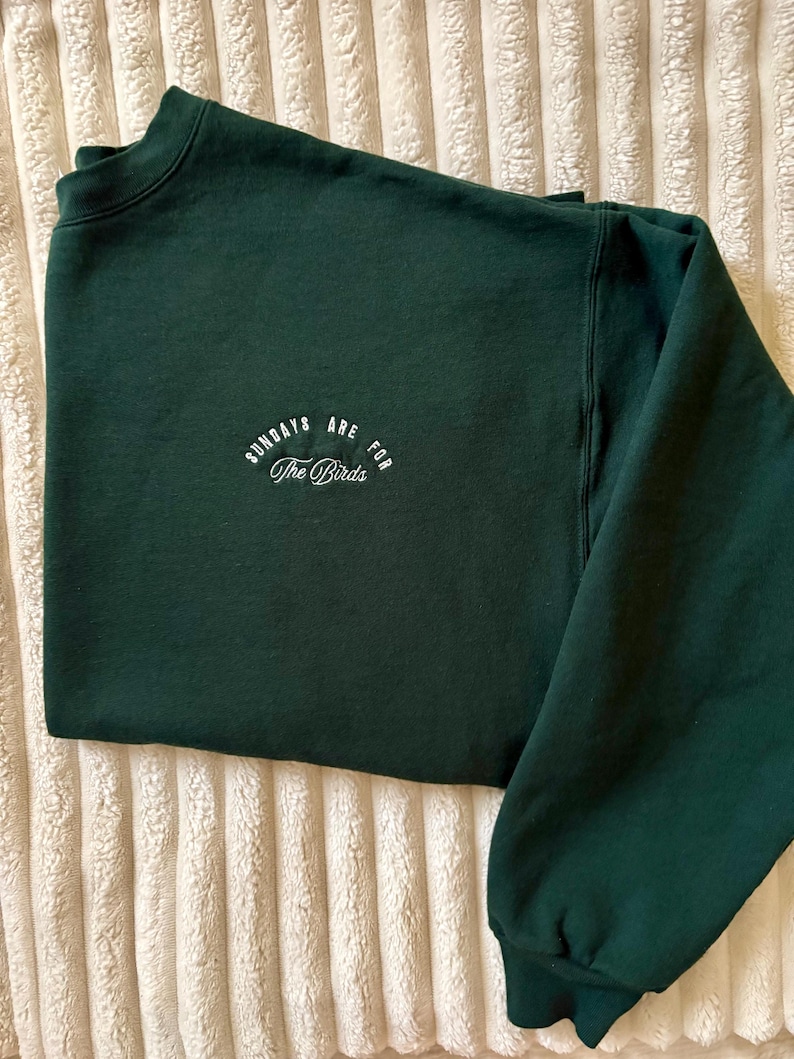 May include: A dark green sweatshirt with the text "SUNDAYS ARE FOR The Birds" in white script. The sweatshirt is folded, showing the crew neck and long sleeves. The fabric appears soft and comfortable.