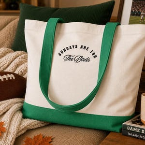 May include: A cream-colored canvas tote bag with green straps and a green bottom panel. The front of the bag features the black text "SUNDAYS ARE FOR The Birds" printed in a combination of sans-serif and script fonts centered on the upper portion.