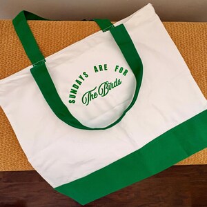 May include: A white canvas tote bag featuring green fabric handles and a green base panel. The front of the bag displays the green printed text "SUNDAYS ARE FOR The Birds" in a combination of block lettering and a cursive script font style.