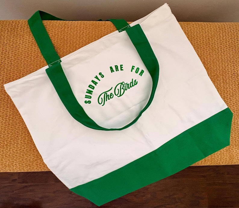 May include: A white canvas tote bag featuring green fabric handles and a green base panel. The front of the bag displays the green printed text "SUNDAYS ARE FOR The Birds" in a combination of block lettering and a cursive script font style.