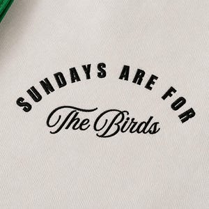 May include: A white fabric garment featuring black embroidered text that reads SUNDAYS ARE FOR in a bold sans-serif font, with the words The Birds written below in a black cursive script. A green fabric edge is visible in the top left corner.