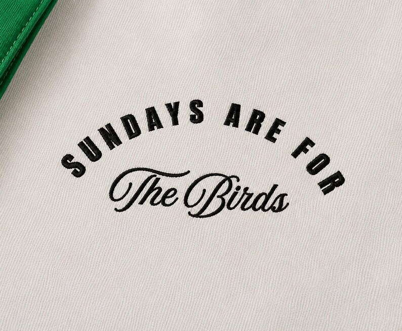 May include: A white fabric garment featuring black embroidered text that reads SUNDAYS ARE FOR in a bold sans-serif font, with the words The Birds written below in a black cursive script. A green fabric edge is visible in the top left corner.
