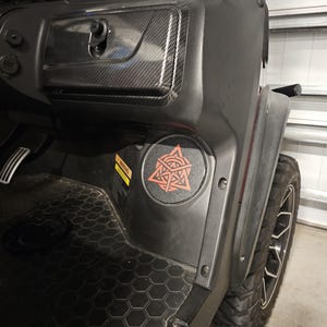 May include: A black vehicle interior dashboard featuring a carbon fiber textured glove box, a circular speaker cover with a red geometric emblem, a yellow warning label, and a black floor mat with a hexagonal pattern near a rugged tire.