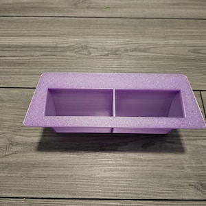 May include: A rectangular lavender plastic container featuring two separate open compartments. The object has a flat, wide rim surrounding the two hollow sections, with a textured surface finish visible across the entire structure.
