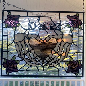 May include: Stained glass panel with a heart formed by skeleton hands. The panel includes the text "10.30.23" and is decorated with purple roses. The artwork is framed in black and hangs from a chain.
