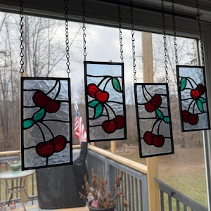 May include: Four rectangular stained glass panels hang from chains. Each panel features a cluster of red cherries with green leaves and black stems. The panels are framed in black and hang in front of a window.