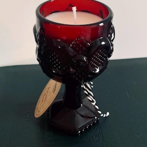May include: A red glass candle in a decorative goblet. The candle has white wax and a single wick. The goblet features ornate detailing and a stem. A small tag is attached with a white and black string.