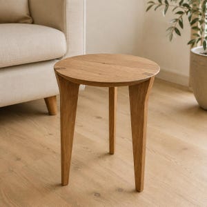 Könnte beinhalten: Ein runder Beistelltisch aus Holz mit einer natürlichen hellbraunen Oberfläche, bestehend aus einer glatten runden Tischplatte und drei konisch zulaufenden Beinen, die von der Mitte nach außen verlaufen. Die Holzmaserung ist sichtbar.