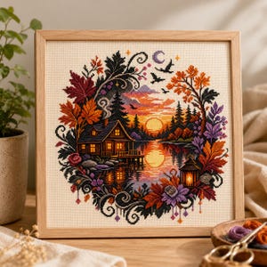 May include: A square cross-stitch embroidery in a light wood frame depicting a cabin by a lake at sunset. The scene is framed by colourful autumn leaves in shades of red, orange, and purple, with a glowing lantern, birds, and a crescent moon.