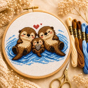 Könnte beinhalten: Ein runder Stickrahmen aus Holz mit einem Kreuzstichmuster von drei braunen Ottern, die in blauem Wasser schwimmen. Die zwei größeren Otter halten einen kleineren Otter zwischen sich, darüber ist ein rotes Herz gestickt.