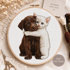Può includere: Un telaio da ricamo a punto croce con un cucciolo marrone e un gattino bianco che si abbracciano. Il testo "Stitched with love" è su una piccola carta. Il disegno è su tessuto bianco e il telaio è in legno. È visibile anche la scritta "PDF PATTERN".