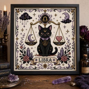 May include: A framed cross-stitch embroidery featuring a black cat sitting beneath a golden scale of justice. The design includes a crescent moon, a heart, purple crystals, roses, and the text LIBRA, all set against a cream-colored fabric background.
