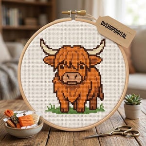 Puede incluir: Un bastidor de bordado circular de madera con un patrón de punto de cruz de una vaca Highland marrón sobre hierba verde. Una etiqueta color canela con la inscripción DVSHOPDIGITAL está sujeta a la parte superior de la tela Aida blanca.