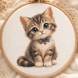 May include: A cross-stitch embroidery of a brown tabby kitten with large blue eyes, wearing a small blue bow tie with a heart charm, set within a round wooden embroidery hoop on a white fabric background with a visible grid pattern.