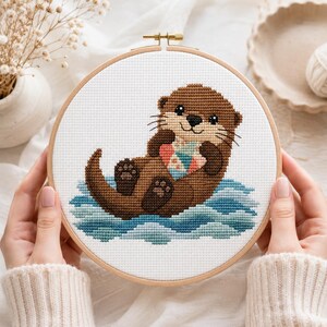 May include: A circular wooden embroidery hoop containing a cross-stitch pattern of a brown otter floating on blue water. The otter is holding a small, multi-colored heart-shaped object against its chest. The design is stitched on white fabric.